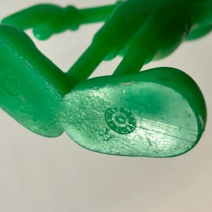 Pu&ograve; includere: Primo piano della parte inferiore di una figura di giocattolo di plastica verde. La parte inferiore del piede ha un'impronta circolare con il testo "ITALY" e "GOOD" all'interno di un cerchio.