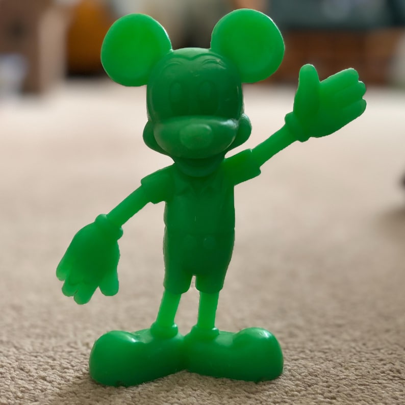 Pu&ograve; includere: Una statuetta di plastica verde di Topolino con il braccio destro alzato e la mano sinistra tesa.
