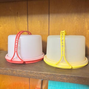 Könnte beinhalten: Zwei Kuchenbehälter aus Kunststoff. Einer ist weiß mit roter Basis und Griff, der andere ist weiß mit gelber Basis und Griff. Beide haben einen transparenten, gewölbten Deckel und stehen auf einem Holzregal.