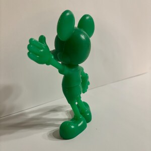 Pu&ograve; includere: Una statuetta di plastica verde di Topolino. La statuetta &egrave; in piedi con il braccio destro teso e il braccio sinistro piegato al gomito. La statuetta &egrave; rivolta a destra.