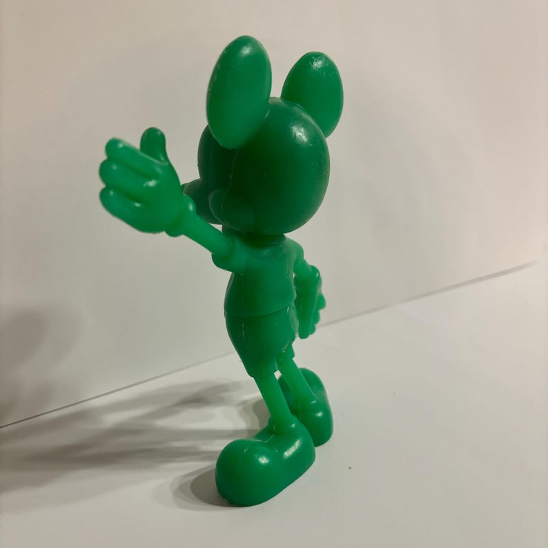 Pu&ograve; includere: Una statuetta di plastica verde di Topolino. La statuetta &egrave; in piedi con il braccio destro teso e il braccio sinistro piegato al gomito. La statuetta &egrave; rivolta a destra.