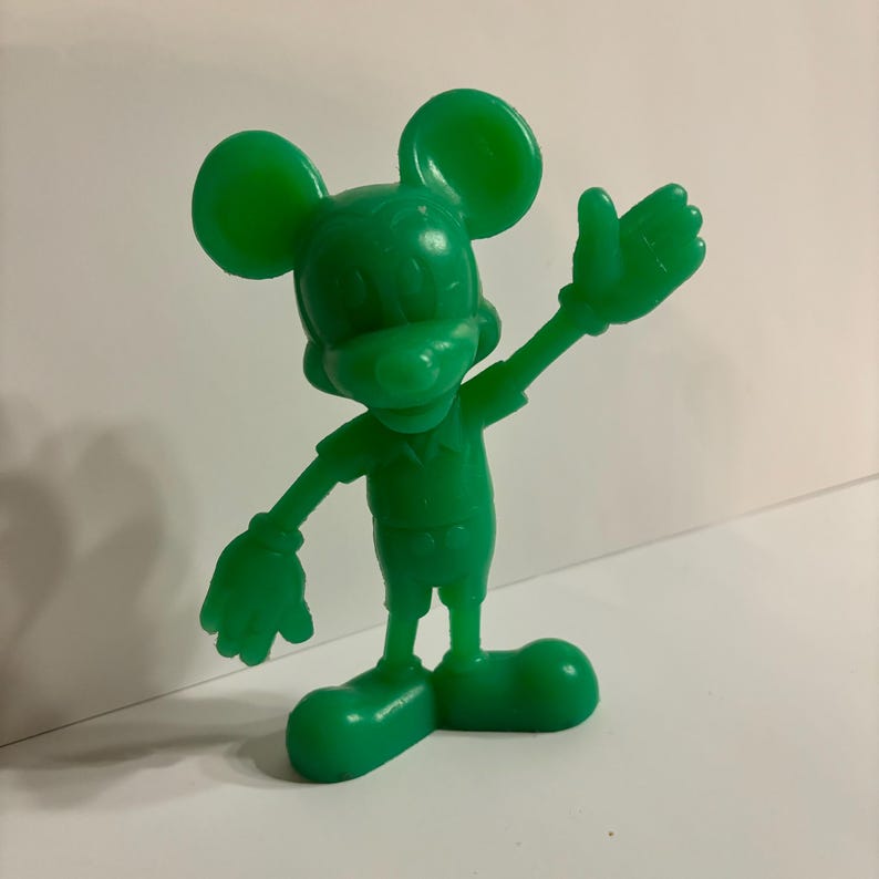 Pu&ograve; includere: Una statuetta di plastica verde di Topolino con il braccio destro alzato in segno di saluto. La statuetta &egrave; in piedi su una superficie bianca.