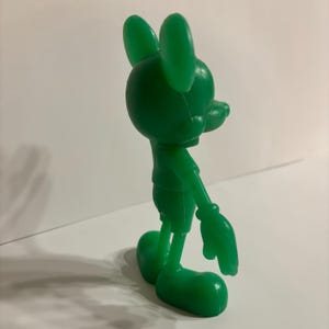 Pu&ograve; includere: Una statuetta di plastica verde di Topolino. La statuetta &egrave; in piedi con il braccio destro teso e il braccio sinistro piegato al gomito. La statuetta &egrave; rivolta a destra.