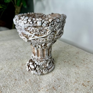 Könnte beinhalten: Ein dekorativer Pflanztopf im Used-Look mit einem Sockel. Der cremefarbene und braune Pflanztopf hat eine strukturierte Oberfläche mit floralen und kieselartigen Details. Geeignet für kleine Pflanzen oder als dekoratives Element.