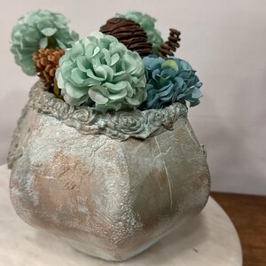 Puede incluir: Un jarrón decorativo y texturizado de color claro con un arreglo floral. El arreglo incluye flores artificiales verde claro y azules, y piñas marrones. El jarrón tiene un borde ornamentado y detallado.