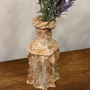 May include: A textured, handmade ceramic vase with a unique, organic shape. The vase has a light brown and cream color palette, with a ruffled design around the base and a flared top. Purple flowers are arranged inside.