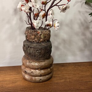 May include: A layered vase with a textured, stone-like finish, displaying branches of white and red flowers. The vase has a unique design with a neutral colour palette, placed on a wooden surface.