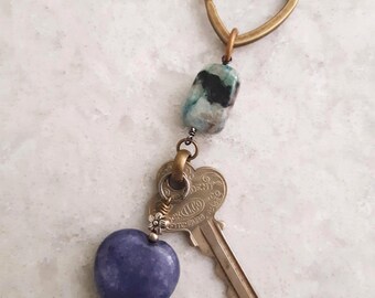 May include: A handmade keychain featuring a heart-shaped brass ring, a blue and black stone, a vintage key, and a heart-shaped blue stone pendant. The key has the text "DENT" and "FITCHBURG" on it.