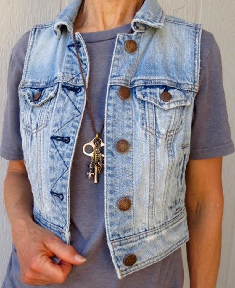 Pu&ograve; includere: Un giubbotto di jeans blu chiaro con una collana di cuoio marrone con un ciondolo a forma di chiave. Il giubbotto ha diverse tasche e bottoni.
