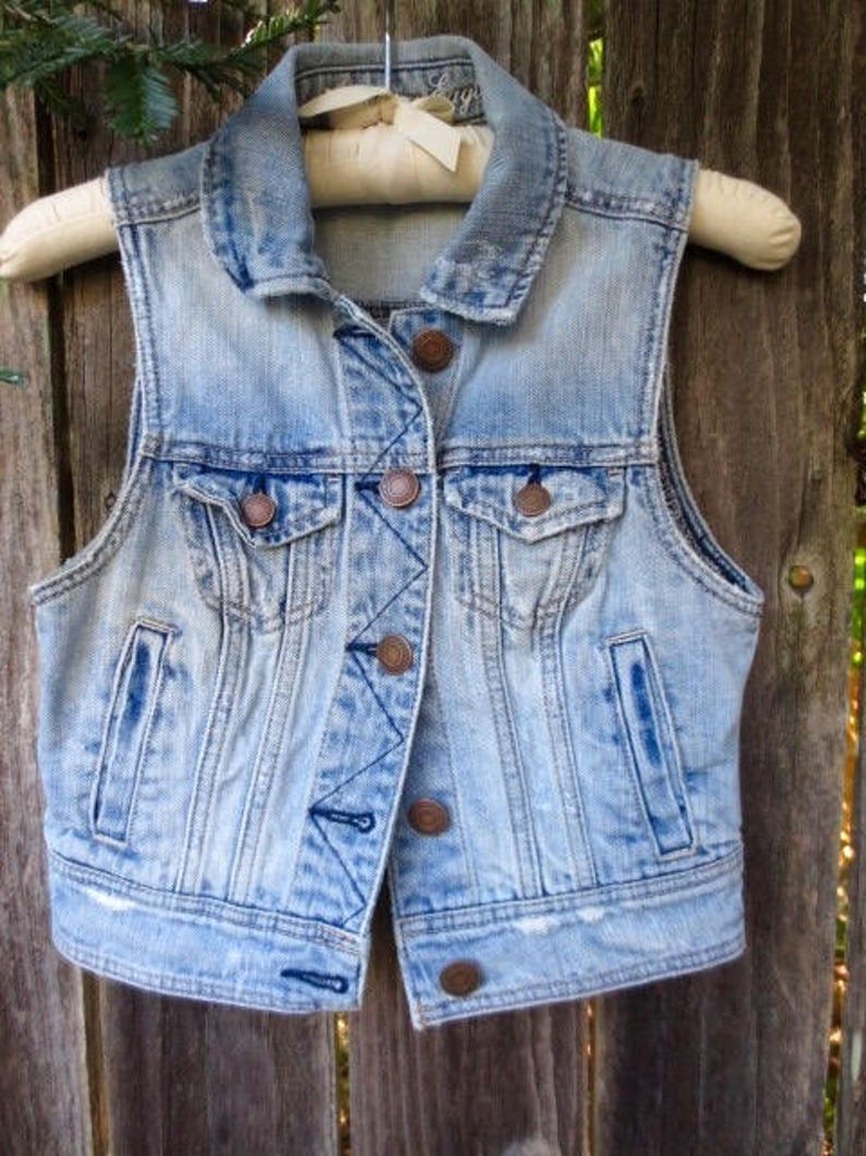 Pu&ograve; includere: Un giubbotto di jeans blu chiaro con colletto classico, chiusura con bottoni e due tasche anteriori. Il giubbotto ha un aspetto vissuto con aree sbiadite e bordi sfilacciati.