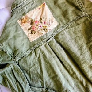 May include: An olive green long-sleeved shirt featuring a rectangular cream-colored fabric patch sewn onto the upper back. The patch displays a detailed floral print with pink, yellow, and white roses surrounded by green leaves and stems.