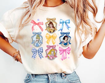 May include: A light beige t-shirt with a graphic print of nine Disney princesses in oval frames with colorful bows. Each princess is wearing a different color dress and has a different colored bow.