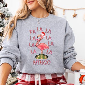 May include: A heather gray crewneck sweatshirt featuring a pink flamingo wearing a Santa hat, surrounded by the text FA LA LA LA LA LA LA LA MINGO. The flamingo is decorated with string lights and stands on a green cactus base.