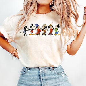 May include: Cream-colored t-shirt featuring a graphic of six Mickey Mouse characters in various outfits. The cartoon characters are in a line, each with a unique costume, including a magician and a cowboy. The shirt is paired with light blue jeans.