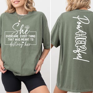 May include: Olive green t-shirt with white text on the front that reads "She overcame everything that was meant to destroy her." The back of the shirt has the word "Powerful" in white. The shirt is a short-sleeved, casual style.