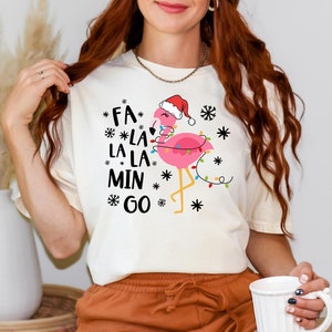 May include: Cream-colored t-shirt featuring a pink flamingo wearing a Santa hat and Christmas lights. The shirt has the text "Fa La La La Min Go" with snowflake accents. The shirt is paired with orange shorts.