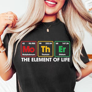 May include: Black t-shirt with a graphic of the periodic table elements Mo, Th, and Er, spelling out "Mother" with the text "The Element of Life" below.