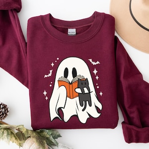 May include: A maroon sweatshirt with a white ghost reading a book with a black cat. The ghost has two eyes and a mouth. The cat is sitting on the book. The background is white with stars and bats.