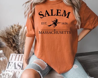 May include: Orange short-sleeved t-shirt with the word "Salem" in black, above "Est. 1626." and "Massachusetts." A witch on a broom is between "Est." and "1626."