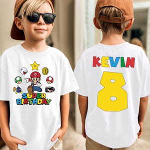 May include: White t-shirt featuring a Super Mario graphic with the text "SUPER BIRTHDAY" in colorful letters. The back of the shirt has the name "KEVIN" and the number "8" in yellow. The shirt is worn by a child.