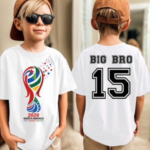 May include: White t-shirt with a colorful graphic of a trophy and the text "2026 NORTH AMERICA USA CANADA MEXICO". The back of the shirt says "BIG BRO 15" in black and white.