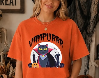 May include: An orange short-sleeve t-shirt featuring a graphic of a black cat dressed as a vampire with a red cape, yellow eyes, and a collar. The text VAMPURRR is printed above the cat, which is set against a white moon, a fence, and tombstones.