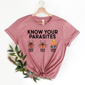 May include: A dusty rose t-shirt with the text "KNOW YOUR PARASITES" above illustrations of a deer tick, dog tick, and "Luna Tick". The shirt is hanging on a wooden hanger.