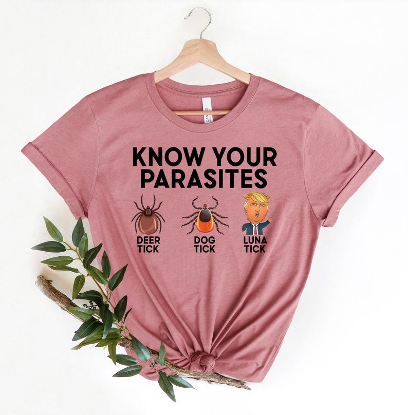 May include: A dusty rose t-shirt with the text "KNOW YOUR PARASITES" above illustrations of a deer tick, dog tick, and "Luna Tick". The shirt is hanging on a wooden hanger.