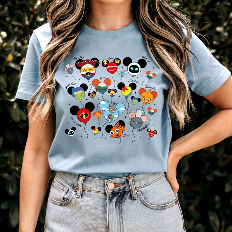 May include: Light blue t-shirt featuring a colorful graphic of Mickey Mouse-shaped balloons. Each balloon has a different character design, with the names of the characters written above. The balloons are connected by strings and float against a light blue background.