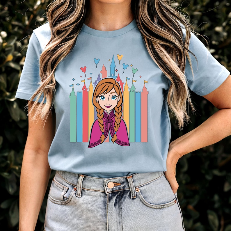 May include: A light blue t-shirt featuring a cartoon image of Anna from Frozen in front of a rainbow-coloured castle with Mickey Mouse balloons.