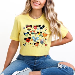 May include: A yellow t-shirt featuring a colorful graphic of character-themed balloons. The balloons have various designs and colors, with black Mickey Mouse ears. The shirt is worn by a person in blue jeans.