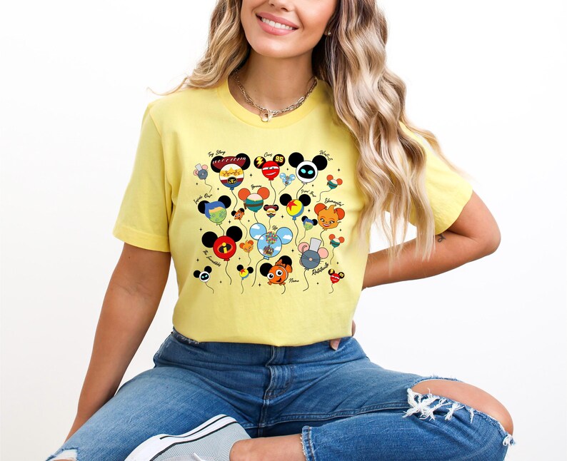 May include: A yellow t-shirt featuring a colorful graphic of character-themed balloons. The balloons have various designs and colors, with black Mickey Mouse ears. The shirt is worn by a person in blue jeans.
