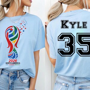 May include: Light blue t-shirt featuring a colorful graphic with the text "2026 NORTH AMERICA USA CANADA MEXICO" on the front. The back of the shirt displays the name "KYLE" above the number "35" in black.