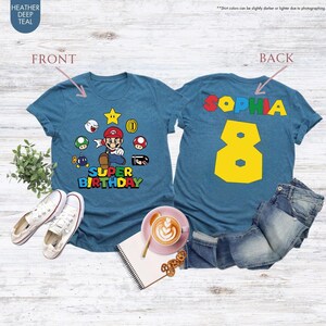 May include: Teal-colored t-shirts with a Super Mario theme. The front shirt features Mario and the words "Super Birthday." The back shirt has the name "Sophia" and the number "8" in yellow. The shirts are displayed with jeans and shoes.