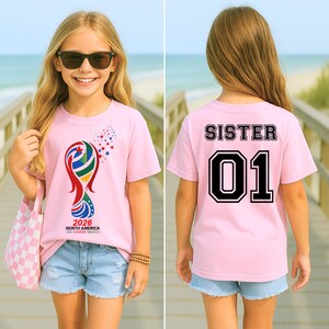 May include: A pink t-shirt with a colorful graphic design and the text "2026 NORTH AMERICA USA CANADA MEXICO" on the front. The back of the shirt has the word "SISTER" above the number "01" in a black outline.