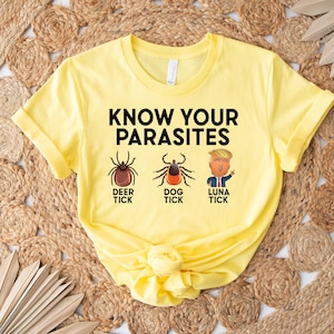 May include: A yellow t-shirt with the text "KNOW YOUR PARASITES" above illustrations of a deer tick, dog tick, and a cartoon of a person. The shirt is folded at the bottom.