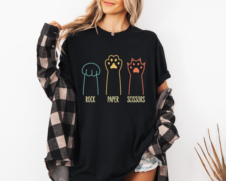 K&ouml;nnte beinhalten: Schwarzes T-Shirt mit einem Grafikdruck von drei Katzenpfoten, die Stein, Papier, Schere darstellen. Die Pfoten sind in verschiedenen Farben umrandet und die Worte "Stein, Papier, Schere" stehen darunter.