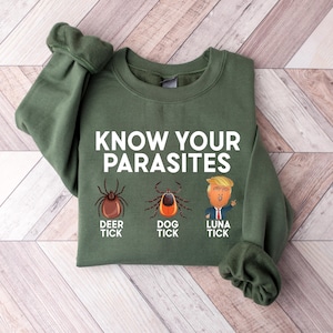 May include: An olive green sweatshirt with the text "KNOW YOUR PARASITES" in white. Below the text are illustrations of a deer tick, a dog tick, and a cartoon of a person with blonde hair. The sweatshirt is made of a soft material.