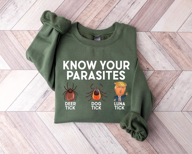 May include: An olive green sweatshirt with the text "KNOW YOUR PARASITES" in white. Below the text are illustrations of a deer tick, a dog tick, and a cartoon of a person with blonde hair. The sweatshirt is made of a soft material.