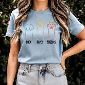 K&ouml;nnte beinhalten: Ein hellblaues T-Shirt mit einer Grafik von Katzenpfoten, die Stein, Papier, Schere spielen. Der Text "Stein, Papier, Schere" ist unter den Pfoten gedruckt.