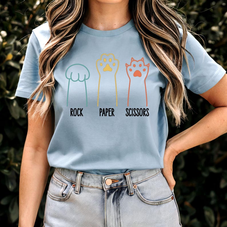 K&ouml;nnte beinhalten: Ein hellblaues T-Shirt mit einer Grafik von Katzenpfoten, die Stein, Papier, Schere spielen. Der Text "Stein, Papier, Schere" ist unter den Pfoten gedruckt.