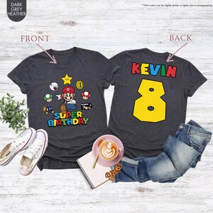 May include: Dark grey heather t-shirts with a Super Mario theme. The front features Mario and other characters with the text "SUPER BIRTHDAY". The back displays the name "KEVIN" and the number "8" in a yellow font.
