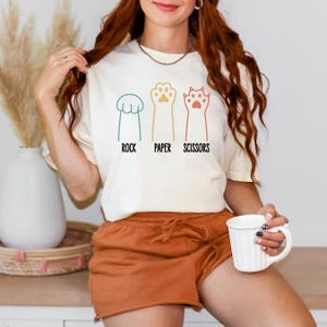K&ouml;nnte beinhalten: Ein wei&szlig;es T-Shirt mit einer Grafik von drei Katzenpfoten, die Stein, Papier, Schere darstellen. Der Text "Stein Papier Schere" ist unter den Pfoten gedruckt.
