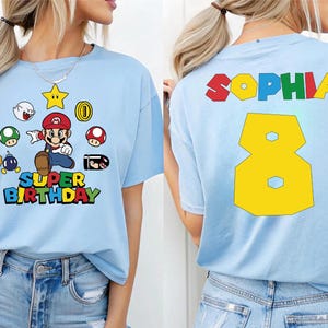 May include: Light blue t-shirt featuring a colorful graphic of a video game character and the words "Super Birthday". The back of the shirt displays the name "Sophia" and the number "8" in yellow.