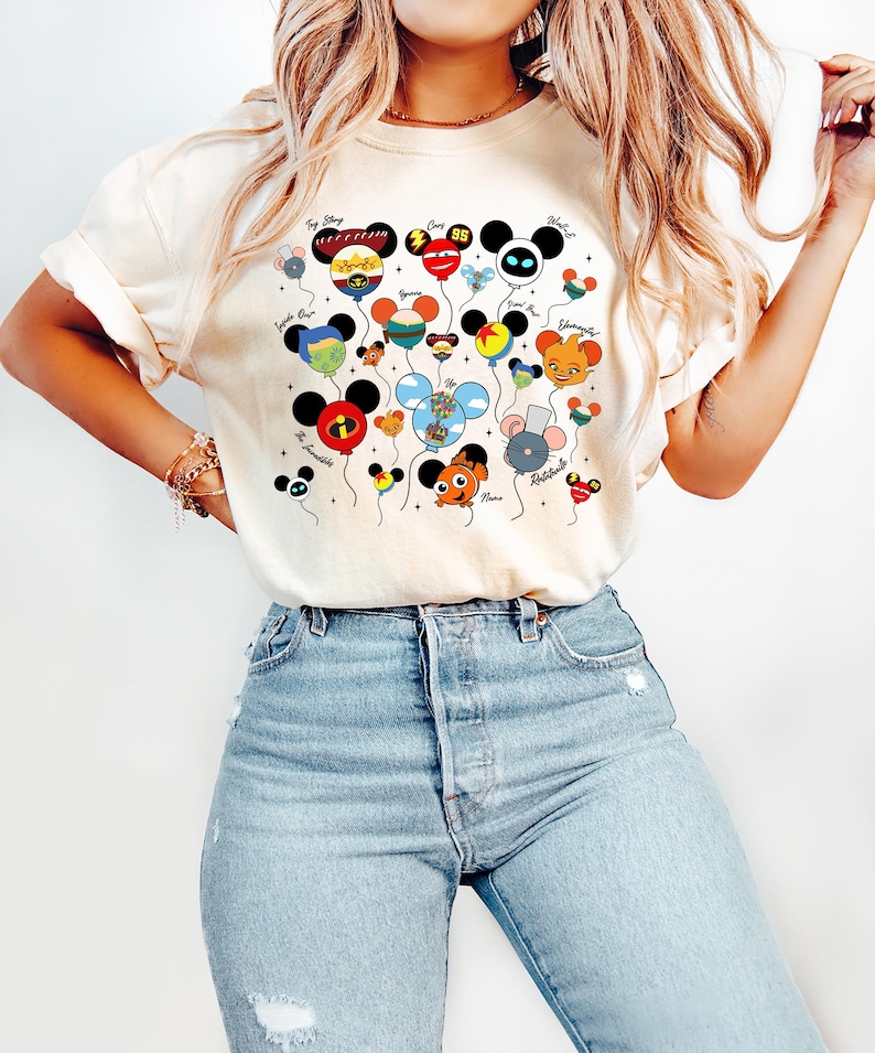 May include: Cream-colored t-shirt featuring a colorful graphic of cartoon character balloons. The balloons are designed with character faces and names, including "Toy Story," "Cars," and "The Incredibles." The shirt is paired with light blue jeans.