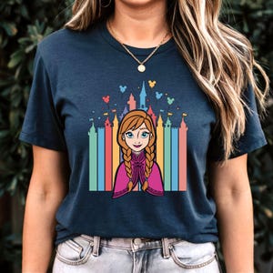 May include: A navy blue t-shirt featuring a colourful graphic of a cartoon princess standing in front of a rainbow-coloured castle. The princess has red hair and is wearing a pink and purple dress. The castle is surrounded by colourful balloons.
