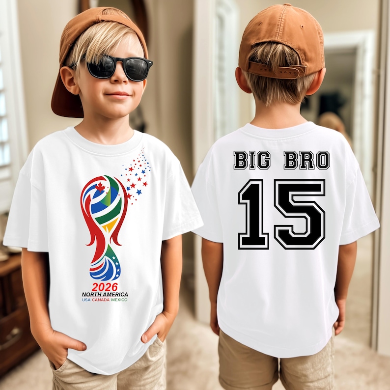 May include: White t-shirt with a colorful graphic of a trophy and the text "2026 NORTH AMERICA USA CANADA MEXICO". The back of the shirt says "BIG BRO 15" in black and white.