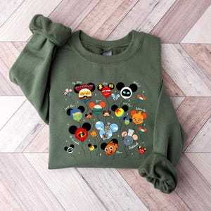 May include: An olive green sweatshirt featuring a colorful graphic of cartoon character-themed balloons. The balloons have Mickey Mouse ears and are labeled with character names. The sweatshirt has long sleeves and a crew neck.