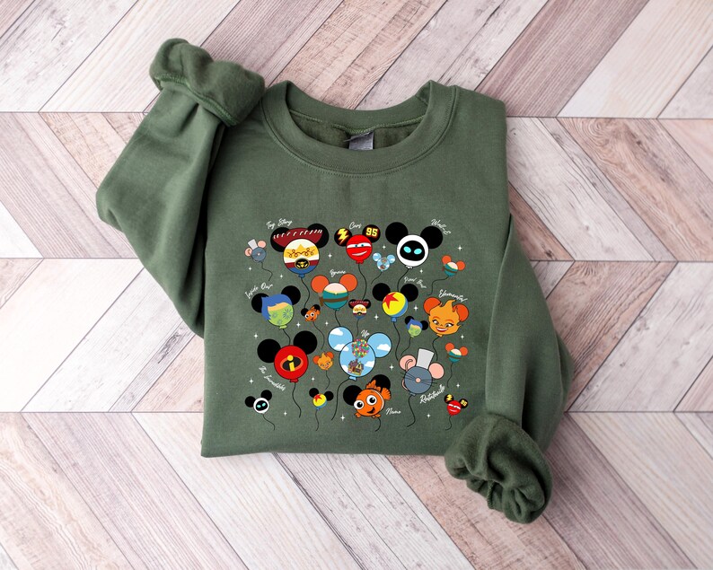 May include: An olive green sweatshirt featuring a colorful graphic of cartoon character-themed balloons. The balloons have Mickey Mouse ears and are labeled with character names. The sweatshirt has long sleeves and a crew neck.