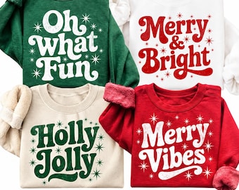 May include: Four crewneck sweatshirts featuring holiday-themed graphics and star accents. The green shirt says Oh what Fun, the white shirt says Merry & Bright, the beige shirt says Holly Jolly, and the red shirt says Merry Vibes.